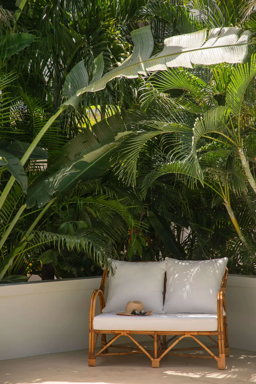 A rattan sofa with white cushions against lush tropical greenery