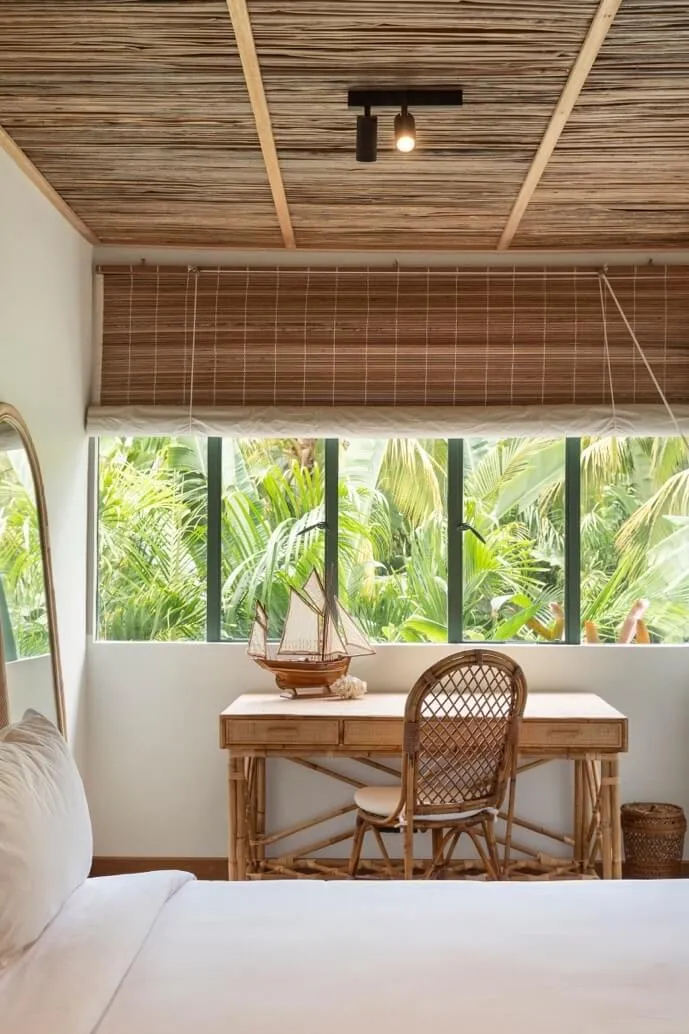 A rattan desk with a model sailing ship, by a bedroom window onto palm trees